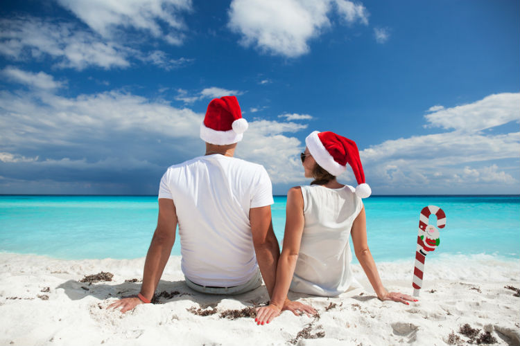 Pärchen von hinten mit Weihnachtsmützen sitzt am Stand vor türkis blauem Meer